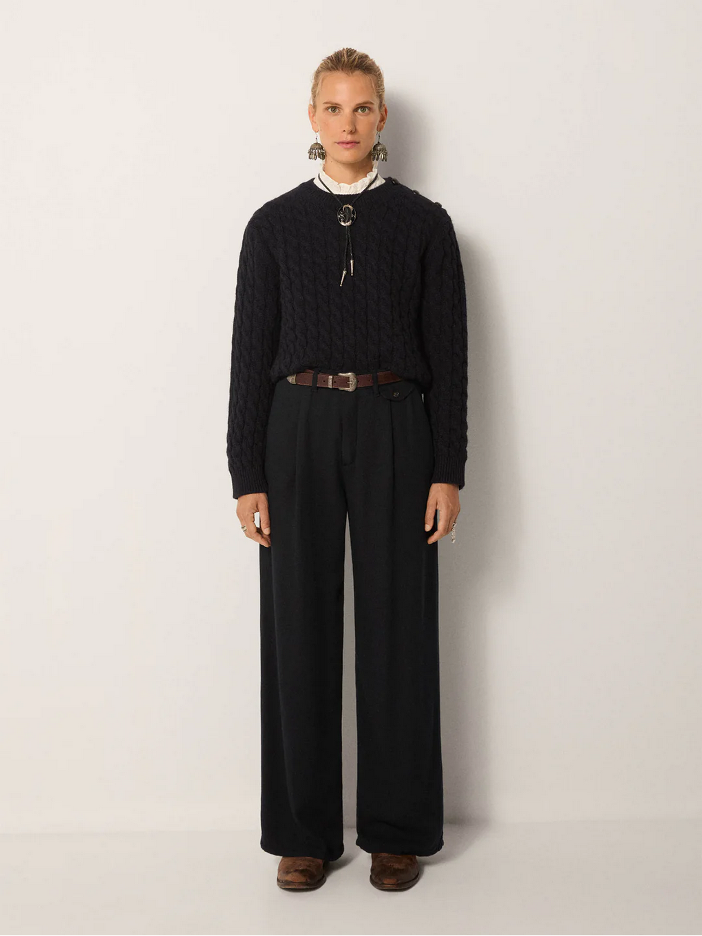 A person stands against a plain background wearing the Keaton Trousers in night blue by Maison Hotel, styled with a cable-knit sweater, brown belt, white collared shirt, and brown shoes.