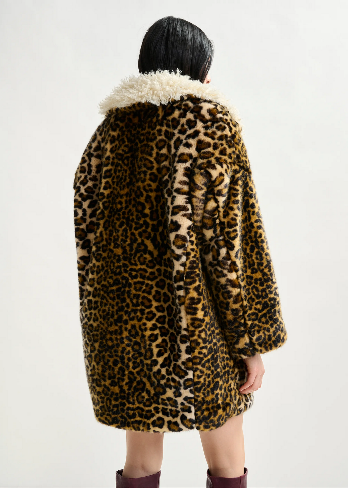 a woman with brown hair wears essentiel antwerp leopard print faux fur coat with cream fluffy collar with red boots against a white background