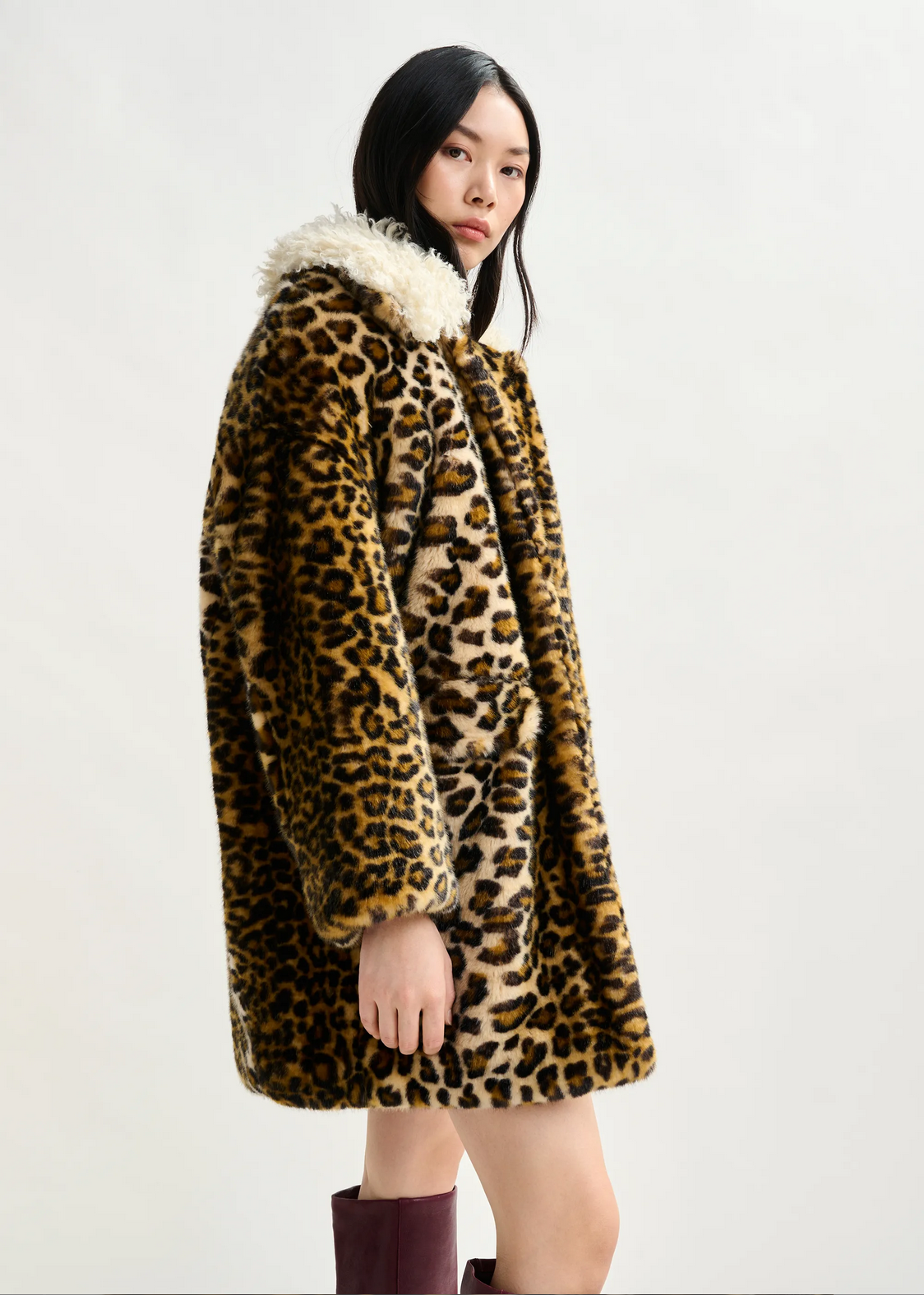 a woman with brown hair wears essentiel antwerp leopard print faux fur coat with cream fluffy collar with red boots against a white background