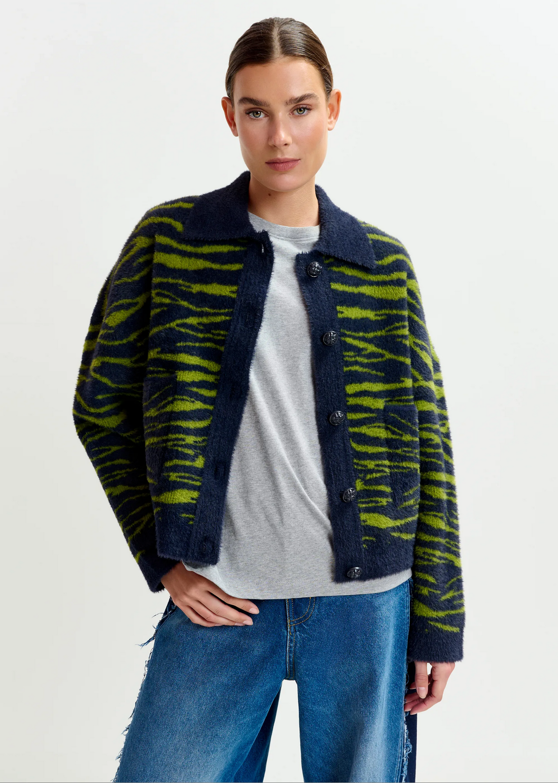 A woman wearing the Essentiel Antwerp Iger Cardigan in dark green and blue over a grey t-shirt and blue jeans stands against a plain white background.