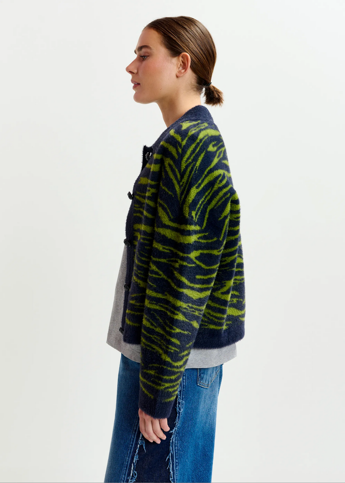 A person stands in profile wearing the Essentiel Antwerp Iger Cardigan in dark green and blue over a gray top and blue jeans, set against a plain light background.