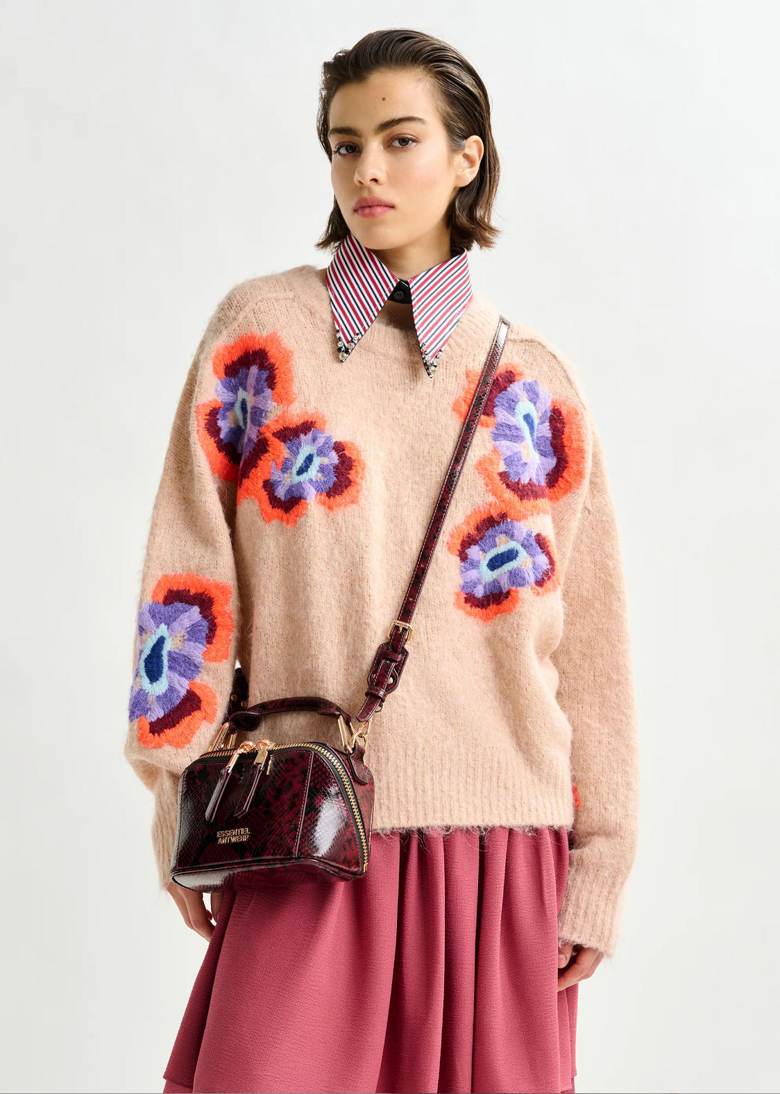A woman wears the Essentiel Antwerp Impossible Jacquard Knit in beige, featuring colorful flower patterns, over a striped collared shirt with a pink skirt and burgundy crossbody bag. She gazes forward with a neutral expression.