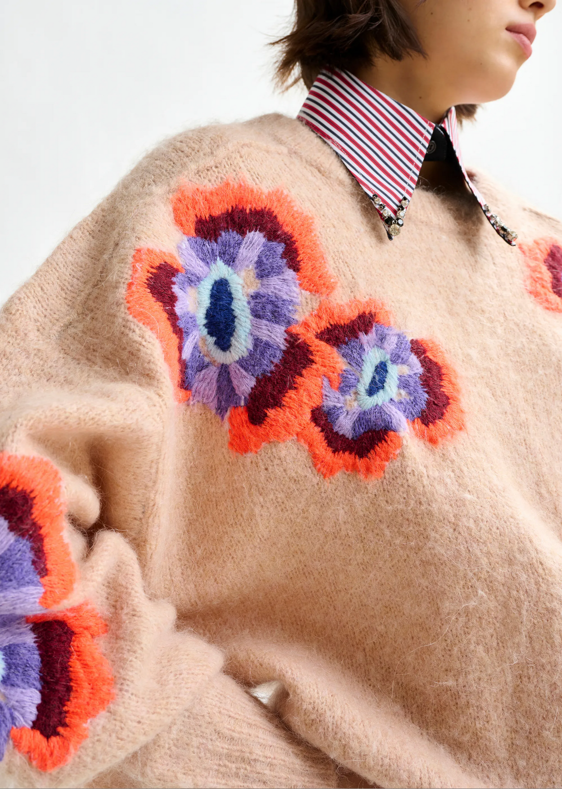 A person wears the Essentiel Antwerp Impossible Jacquard Knit in beige, featuring bold purple, red, and orange floral patterns, layered over a red-and-white striped collared shirt.