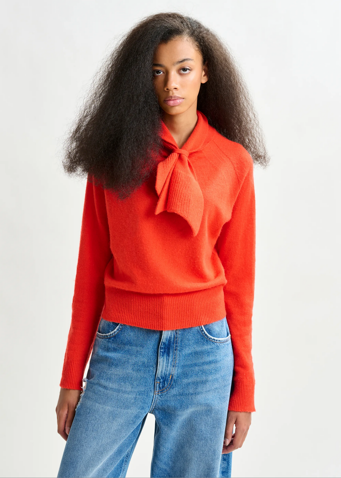 A person wearing the Essentiel Antwerp Invited Cashmere Sweater in vibrant orange, featuring a bow-tie neck detail, paired with blue jeans, standing against a plain light background.