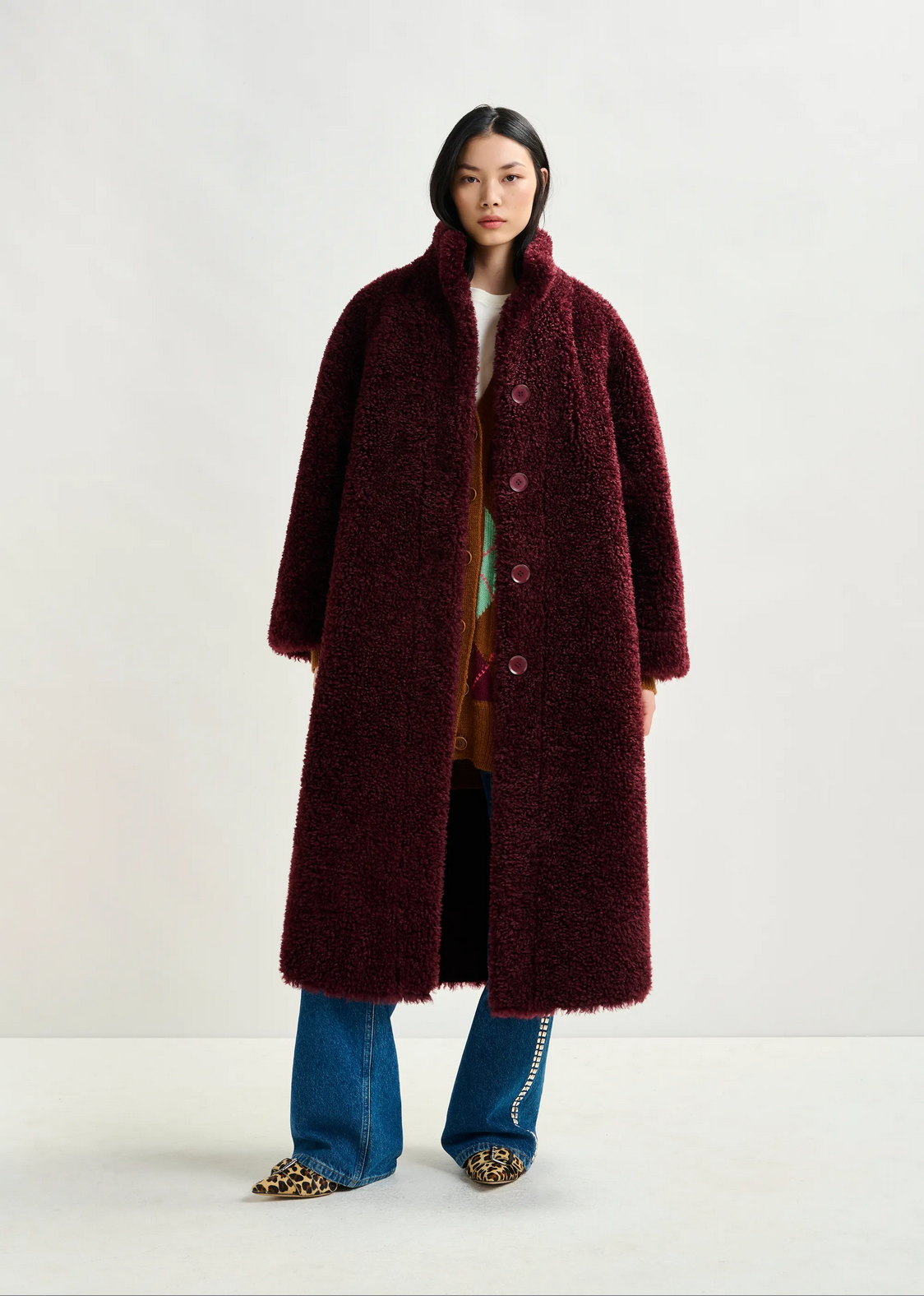 A woman faces forward against a plain light background, wearing the Essentiel Antwerp Ispot Faux Shearling Coat in burgundy over a printed dress and blue jeans, styled with studded leopard print shoes.