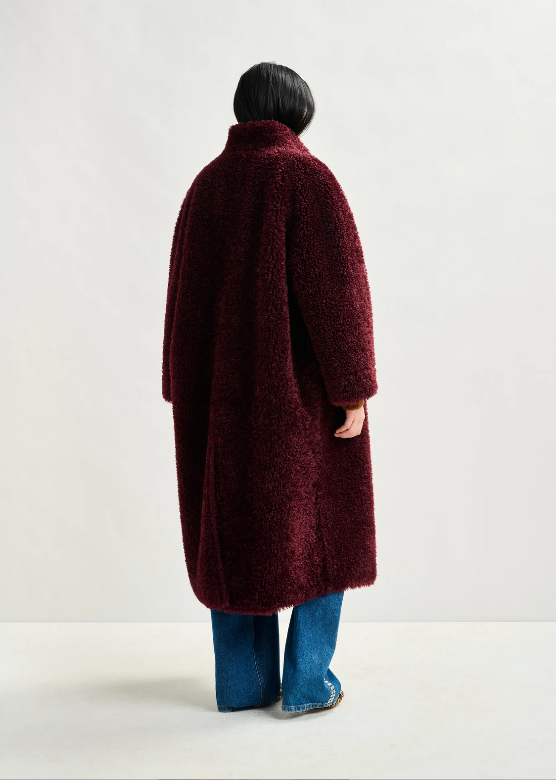 A person wearing the Essentiel Antwerp Ispot Faux Shearling Coat in burgundy and blue jeans stands with their back to the camera against a plain light background.