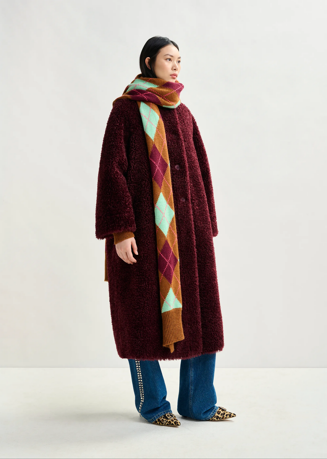 A person wears the Essentiel Antwerp Ispot Faux Shearling Coat in burgundy, paired with blue jeans featuring side embellishments, leopard print shoes, and a colorful argyle scarf against a plain light background.