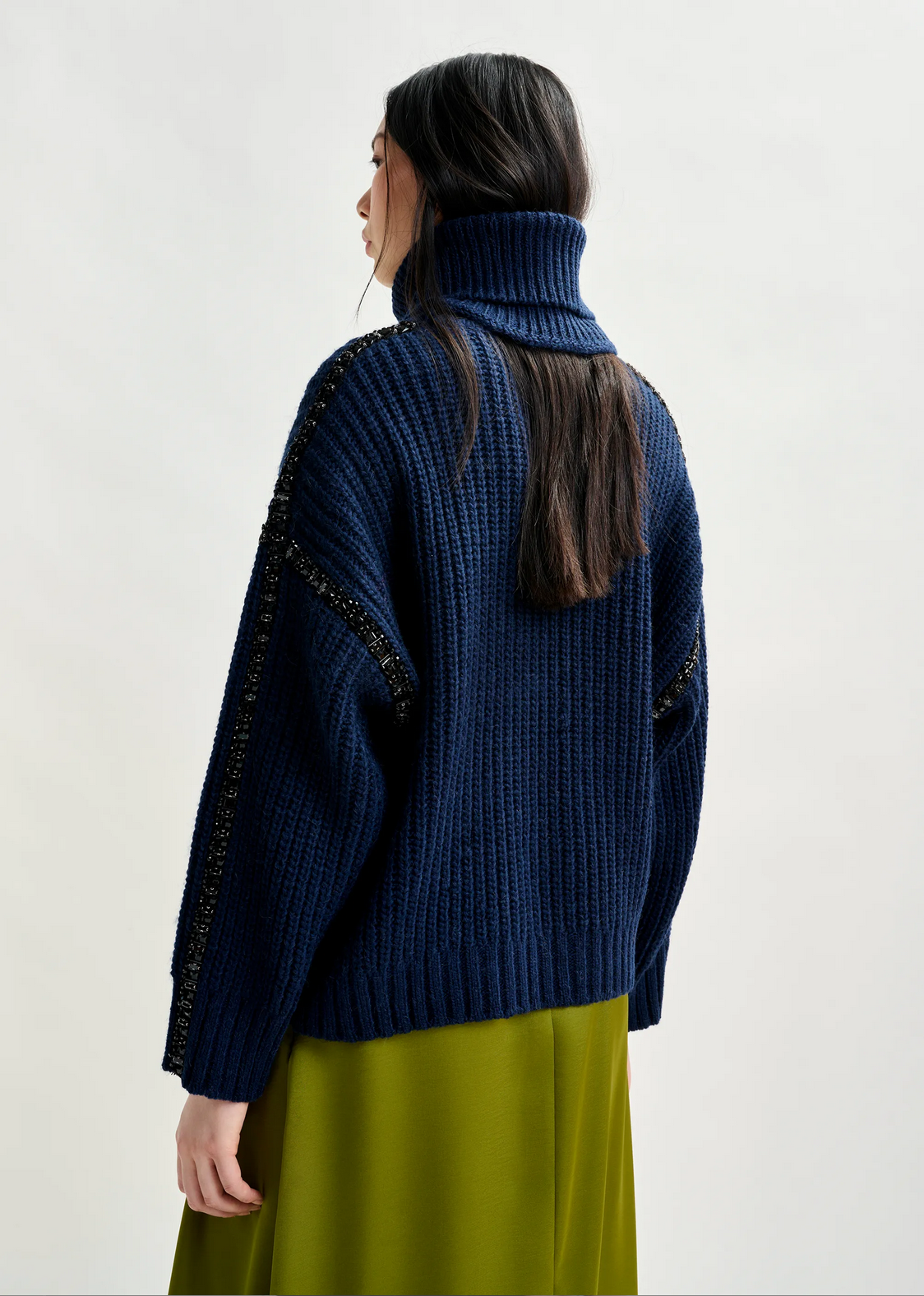 A woman with long dark hair, seen from behind against a plain background, wears the Essentiel Antwerp Indsay Wool Turtleneck in navy and an olive green skirt.