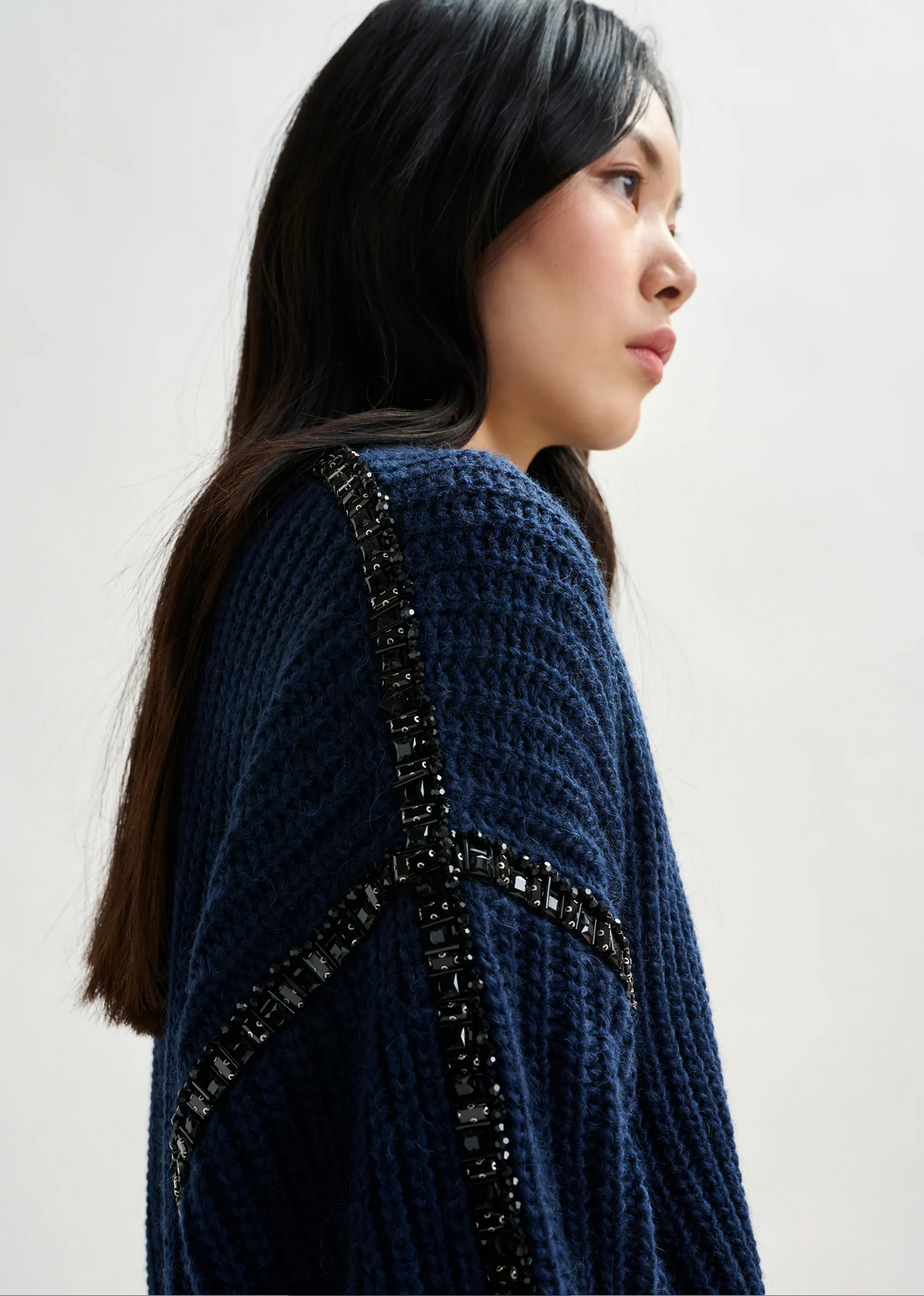 Woman in the Essentiel Antwerp Indsay Wool Turtleneck in navy, featuring textured fabric and black beaded sleeve embellishments, shown in profile against a plain light background.