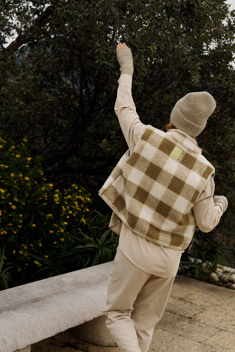 Wearing the Indi & Cold Teddy Plaid Vest in Beige/Olive, a person stands on a stone path by a bench, reaching for tree branches with yellow flowers blooming in the background.