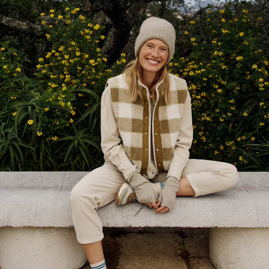 A woman in the Indi & Cold Teddy Plaid Vest in Beige/Olive and a knit hat sits cross-legged on a stone bench, smiling, with green foliage and yellow flowers behind her.