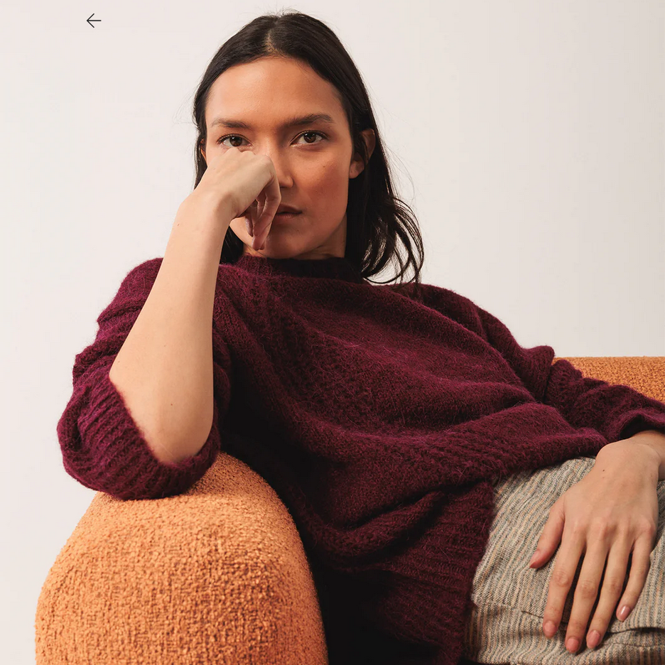 A person with long dark hair sits on an orange textured chair, wearing the Indi & Cold Loose-knit Wool Sweater in burgundy and beige pants, resting one hand on their face while looking at the camera.