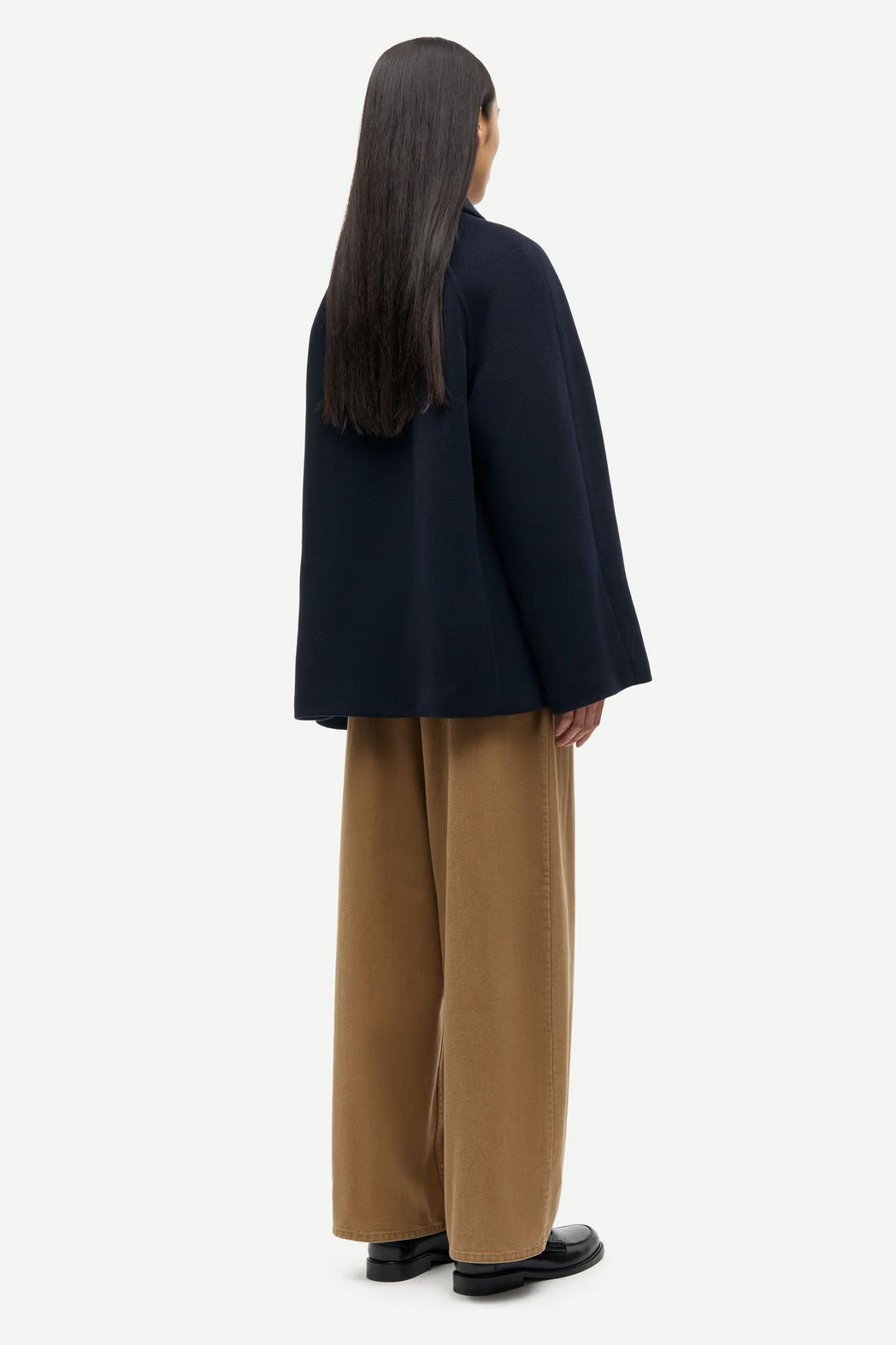 A person stands facing away in the Samsøe Samsøe Alma Jacket - Salute, paired with wide-legged tan pants and black shoes against a plain white background.