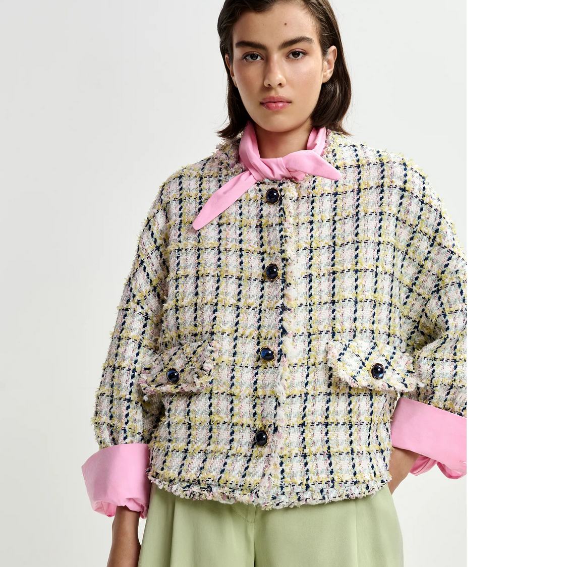A woman wears the Essentiel Antwerp Jayden Boucle Jacket in off white/black/green, featuring black buttons, pink cuffs, frayed trim, and a pink neck scarf. She pairs it with light green pants against a plain background.