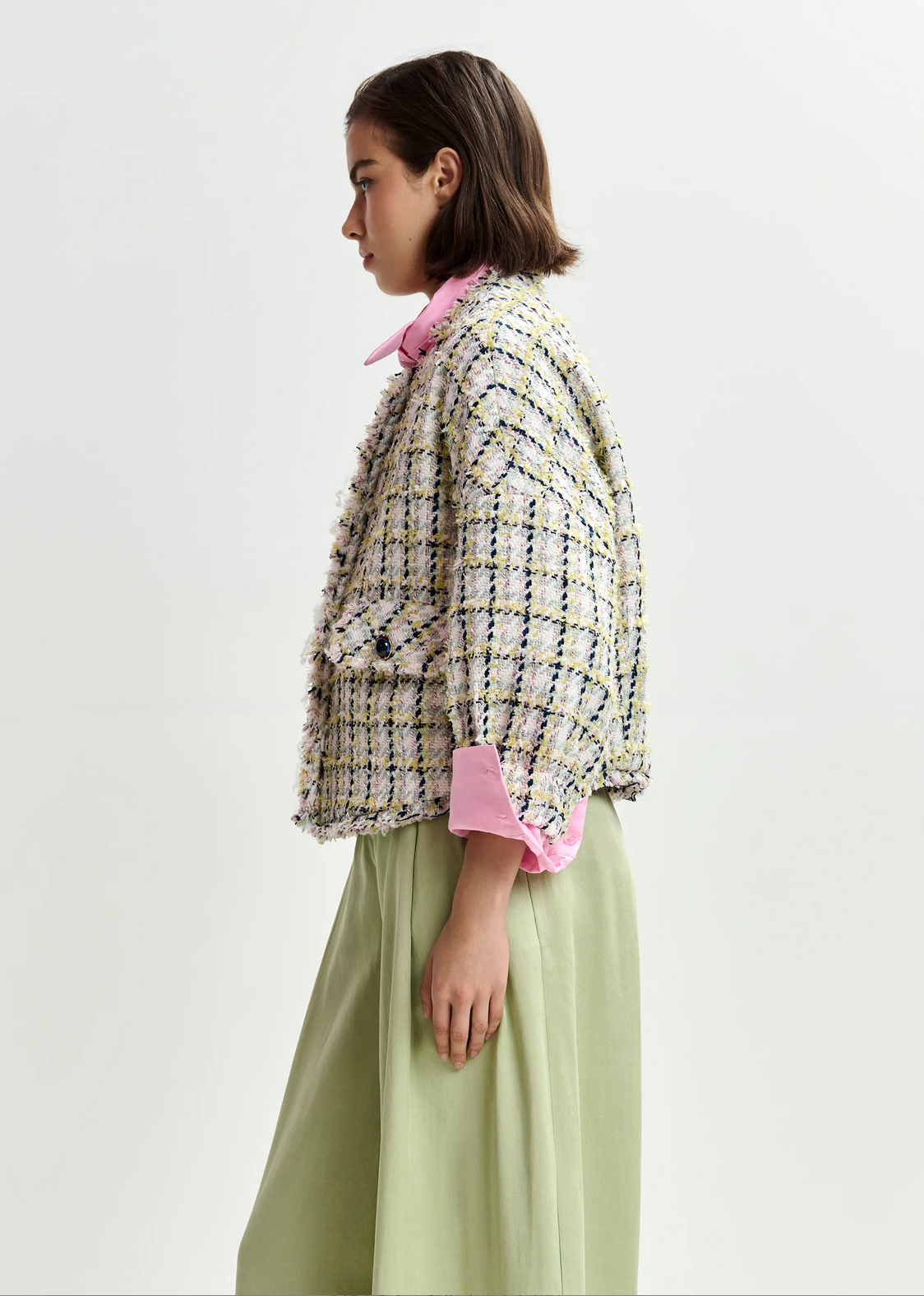 A woman in profile wears the Essentiel Antwerp Jayden Boucle Jacket in Off White/Black/Green, featuring a relaxed fit and frayed trim, layered over a pink collared shirt and light green skirt against a plain backdrop.
