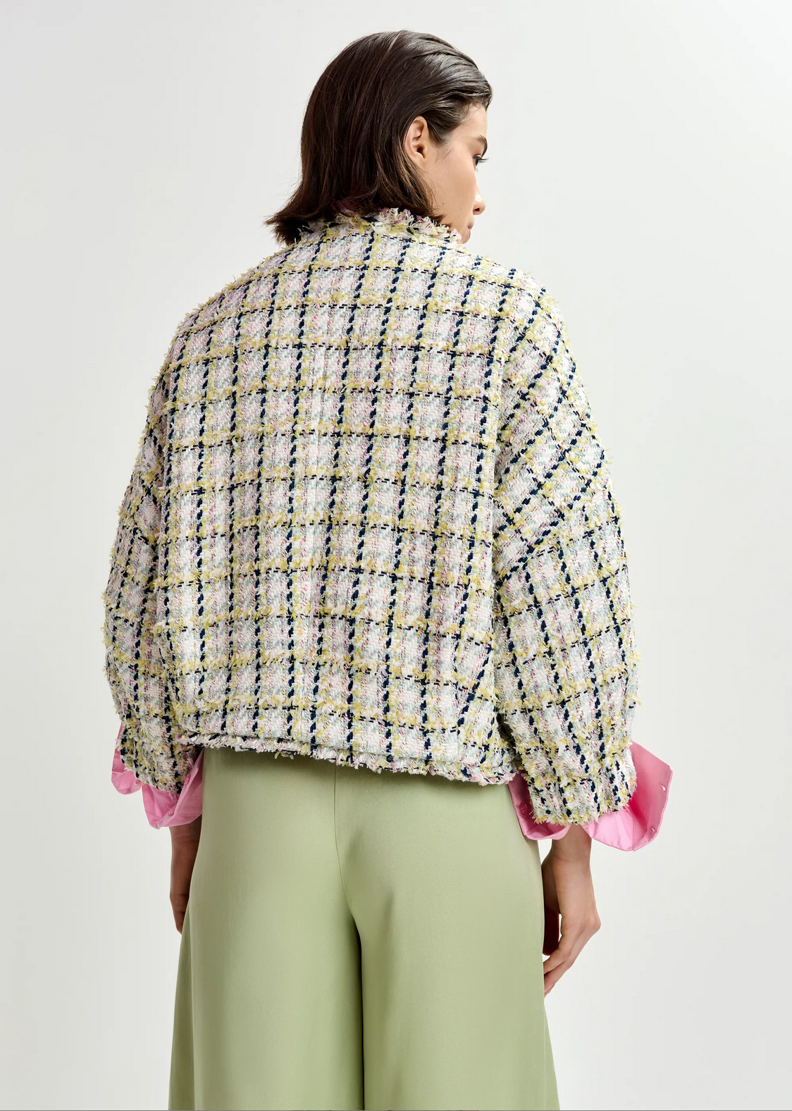 A person stands facing away, wearing the Essentiel Antwerp Jayden Boucle Jacket in Off White/Black/Green with pink shirt cuffs and light green pants against a plain background.