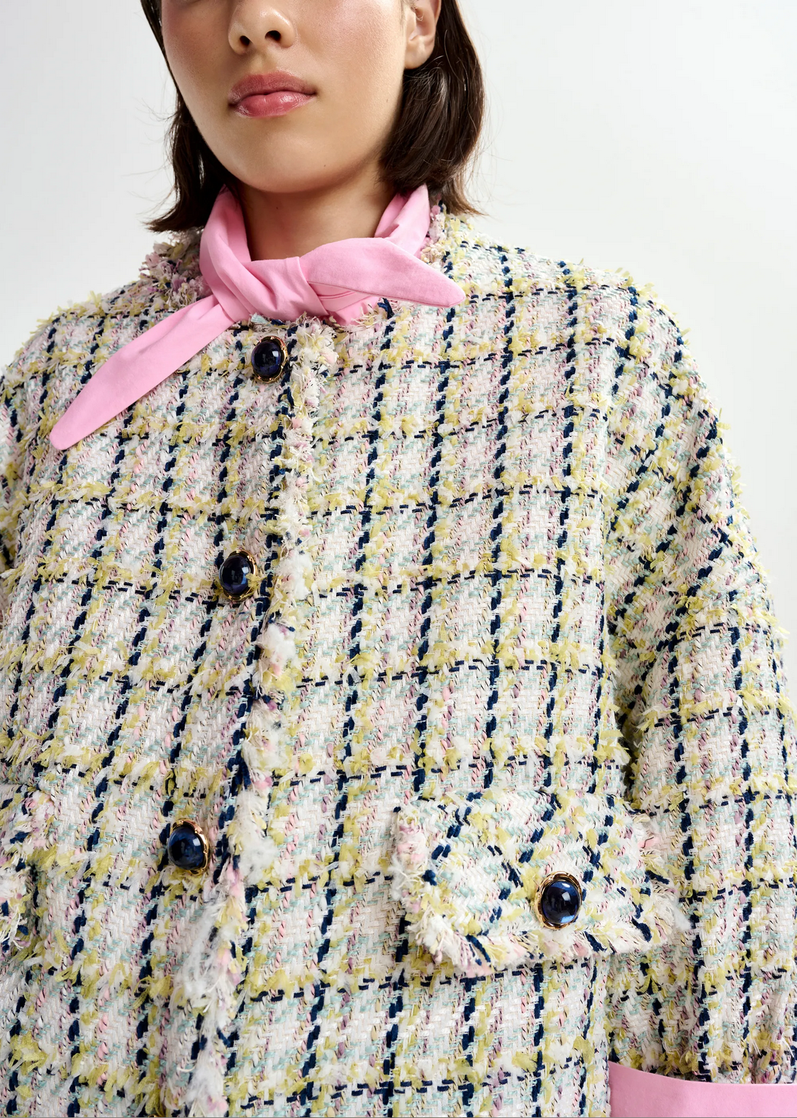 A person wearing the Essentiel Antwerp Jayden Boucle Jacket in Off White/Black/Green, featuring a pastel plaid texture and dark buttons, paired with a light pink neck scarf, shown from shoulders to lips.