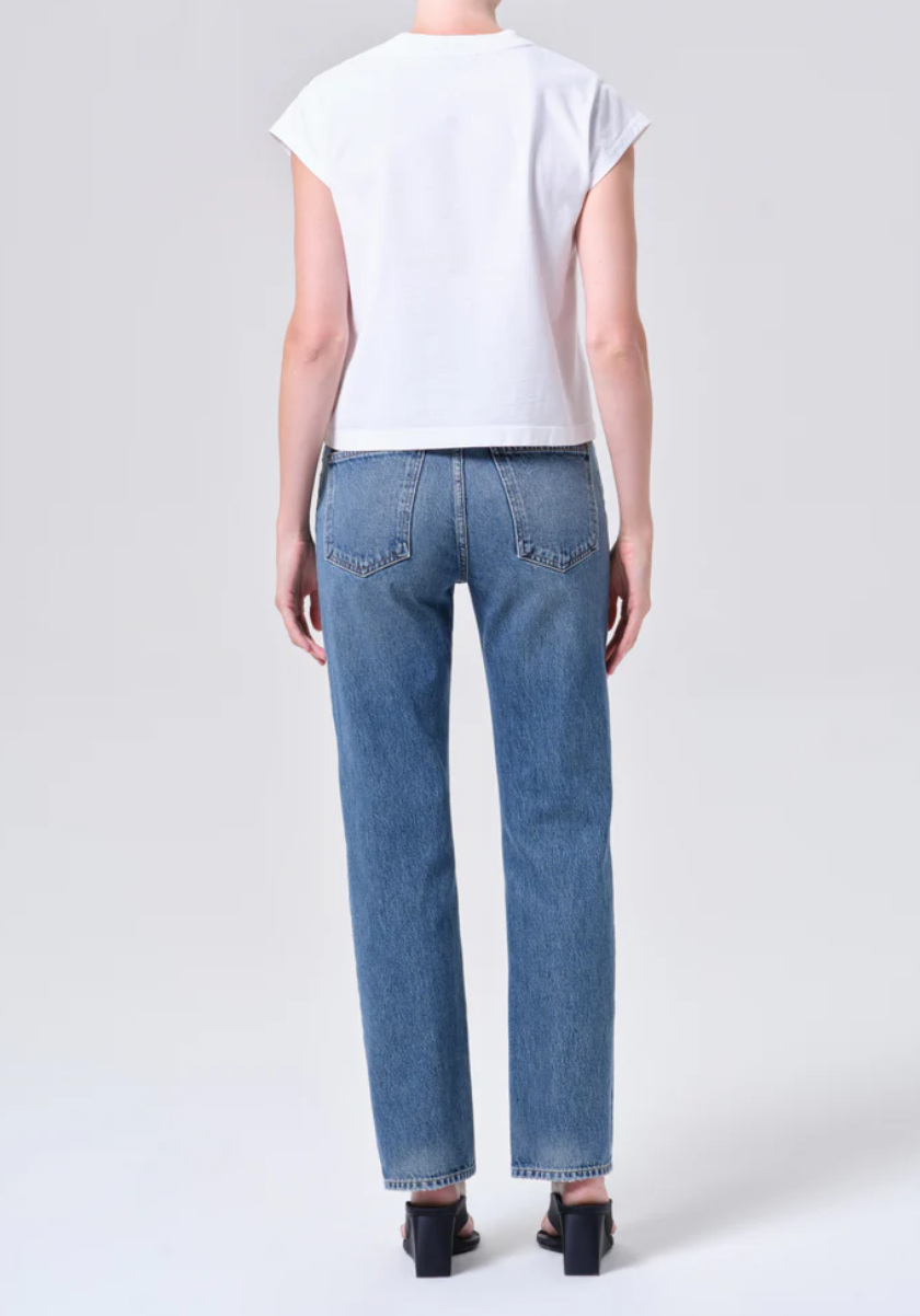 Person standing with back to camera, wearing a white short-sleeve t-shirt, AGOLDE Lana Jean - Liberty in blue regenerative cotton denim, and black heeled sandals against a plain light background.