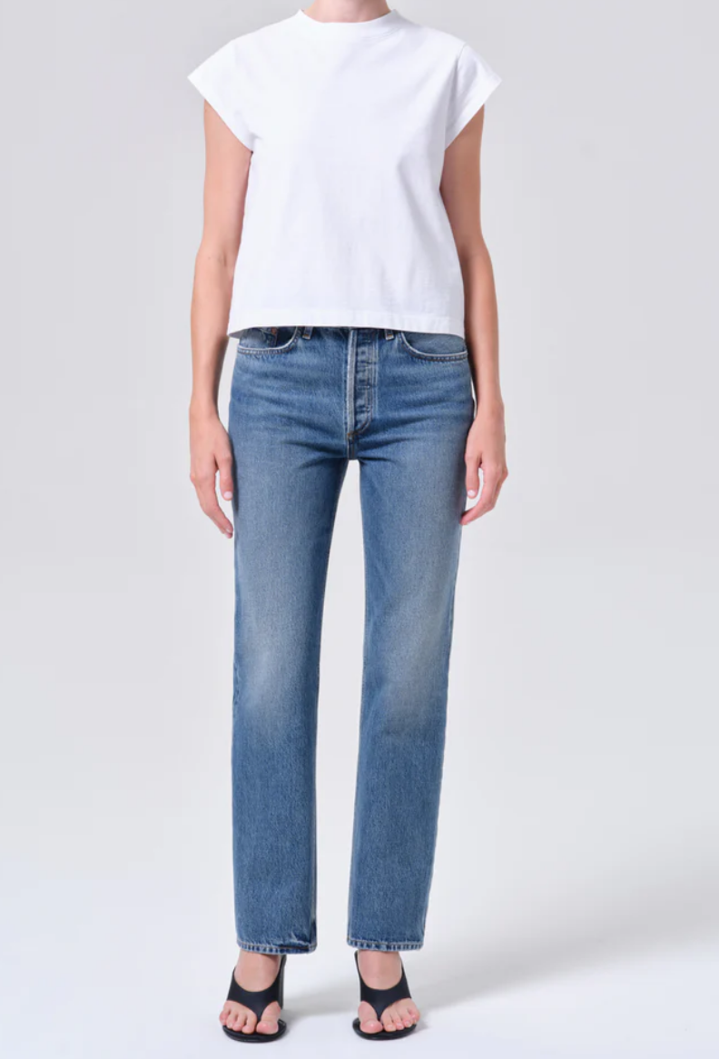 A person wears a white short-sleeve top, AGOLDE Lana Jean - Liberty in blue, and black open-toe heeled sandals while standing against a light gray background.