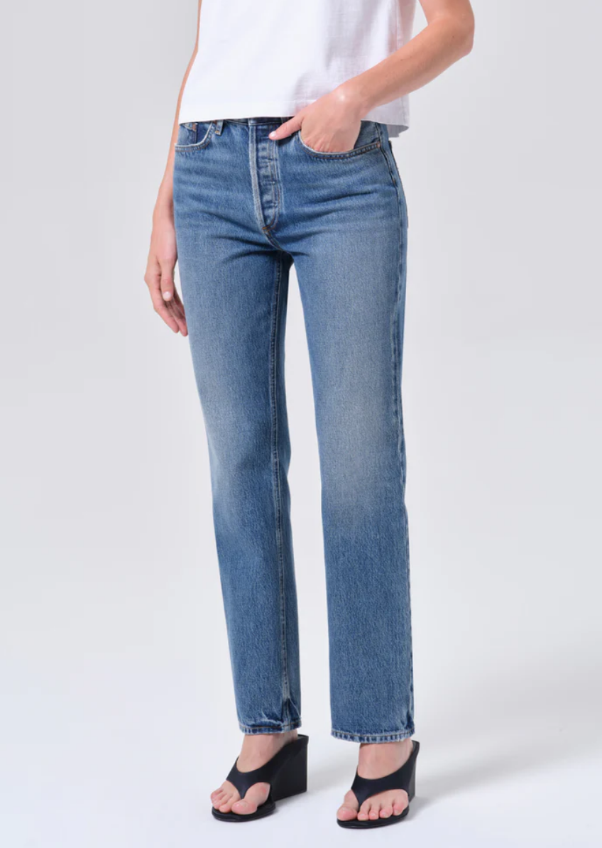 A person wears the AGOLDE Lana Jean - Liberty, blue straight-leg jeans made from regenerative cotton denim, with a white top and black wedge sandals, one hand in a pocket, against a plain background.