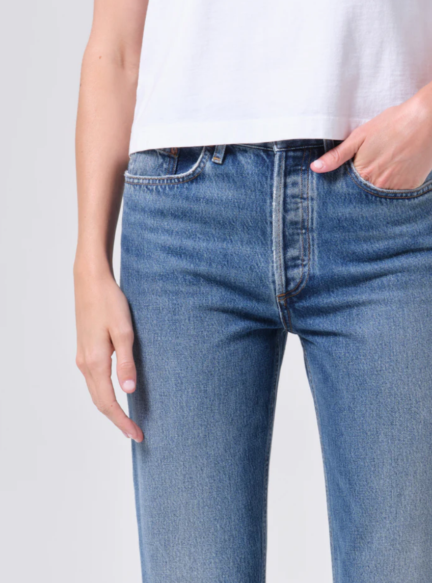 A person wearing AGOLDE's Lana Jean - Liberty, crafted from regenerative cotton denim, and a white shirt stands with one hand in a pocket against a plain background.
