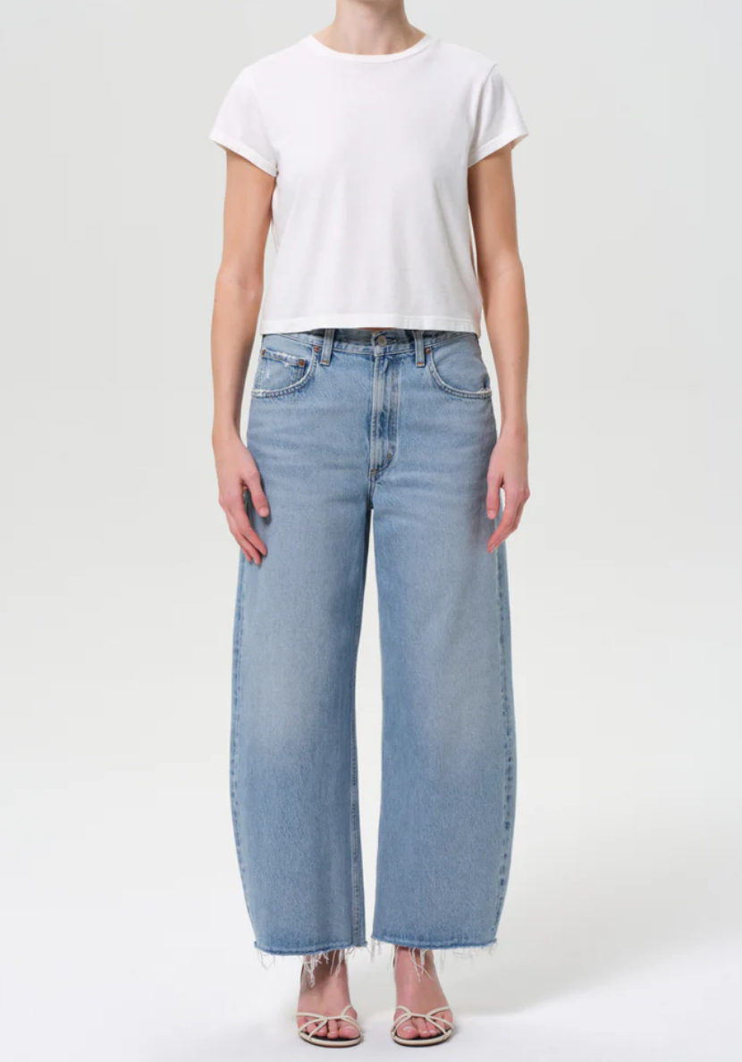 A person wearing a plain white short-sleeve t-shirt, AGOLDE Low Curve Fray - Force jeans with light blue wash and frayed hems, and open-toe heels stands against a plain light background.