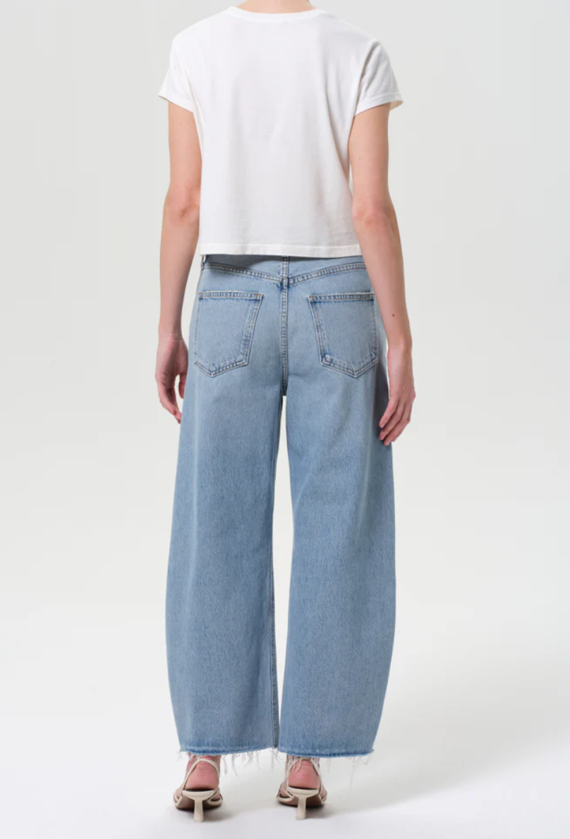 A person faces away, wearing a white short-sleeve crop top and AGOLDE Low Curve Fray - Force light blue high-waisted jeans made from regenerative cotton with a frayed hem, paired with heeled sandals against a plain background.