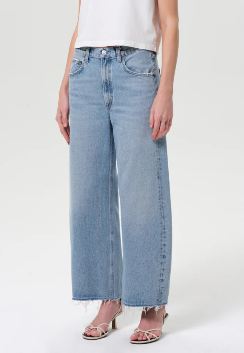 A person in a white sleeveless top, AGOLDE Low Curve Fray - Force jeans in light blue regenerative cotton with frayed hems, and white strappy heels stands against a plain background.