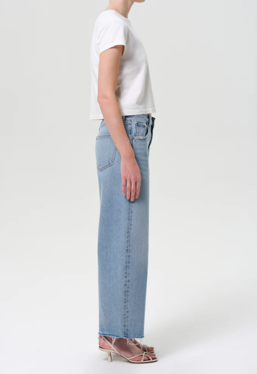 A person stands in profile wearing a white short-sleeve crop top, AGOLDE’s Low Curve Fray - Force jeans in light blue with a frayed hem, and strappy heeled sandals against a plain background.