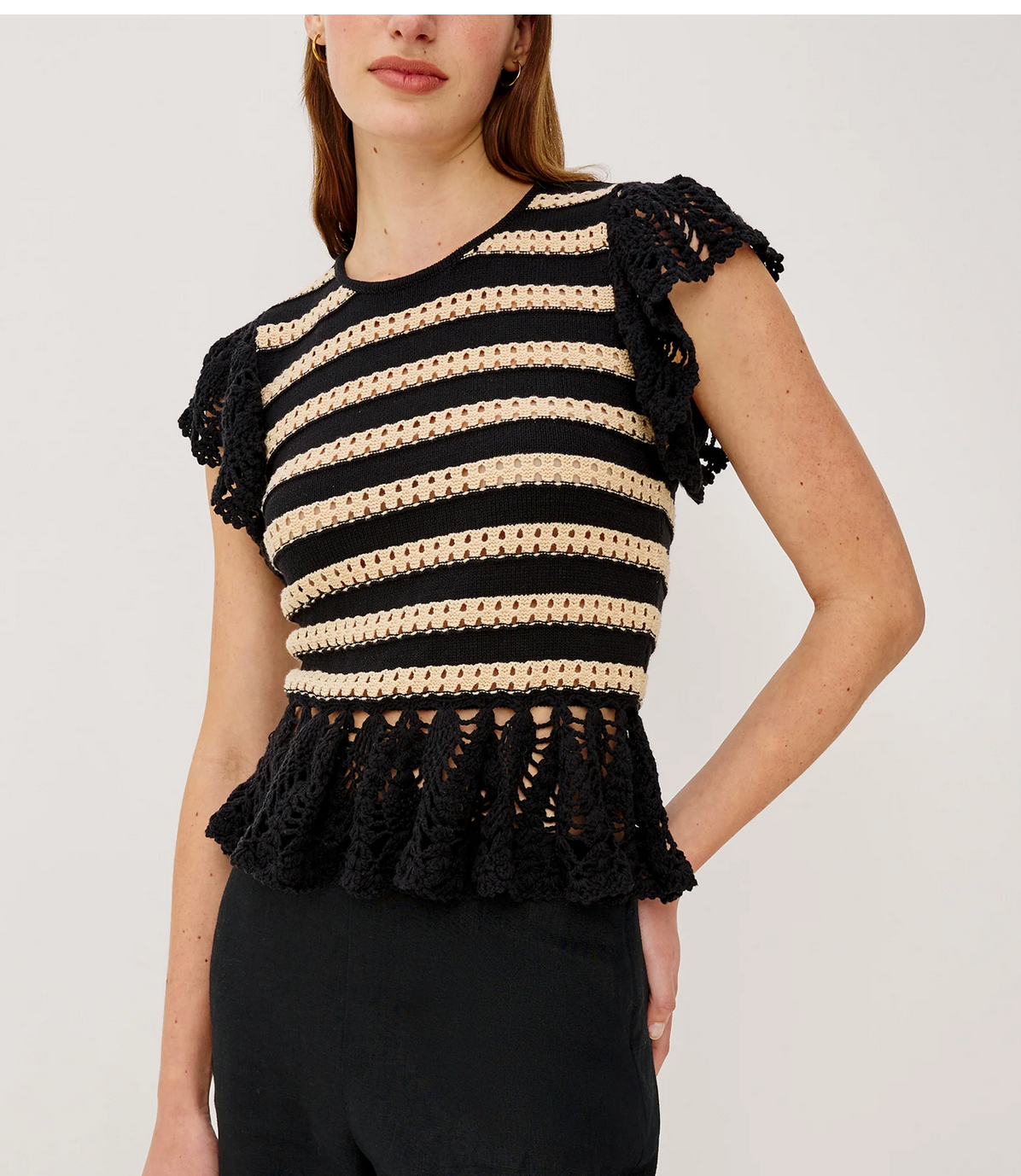 A woman wears the Rails Coen Knit Top in black stripe, featuring short sleeves, nautical-inspired crochet, and a peplum hem, paired with black pants against a plain background.