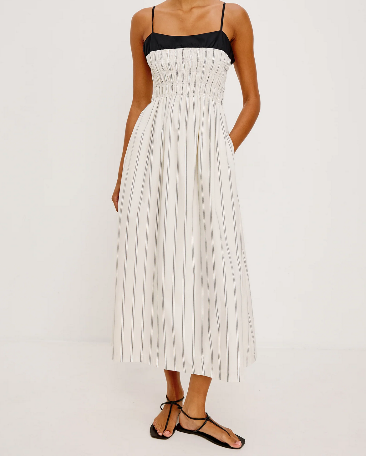 A person wears the Rails Primrose Dress - Ecru Stripe, a sleeveless cotton poplin style with thin straps, black ruched bodice, and white skirt with vertical black stripes, paired with black sandals for a chic look.