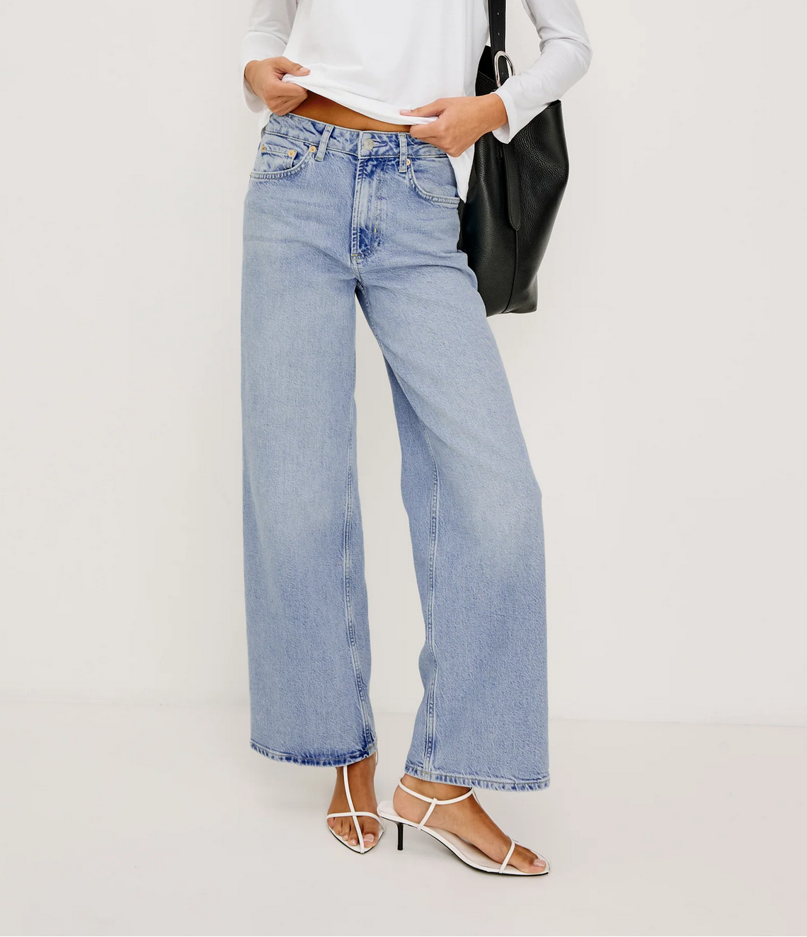A person wearing Rails' Getty Wide Leg - Light Blue jeans, a white long-sleeve top, white heeled sandals, and holding a black bag stands against a plain background.