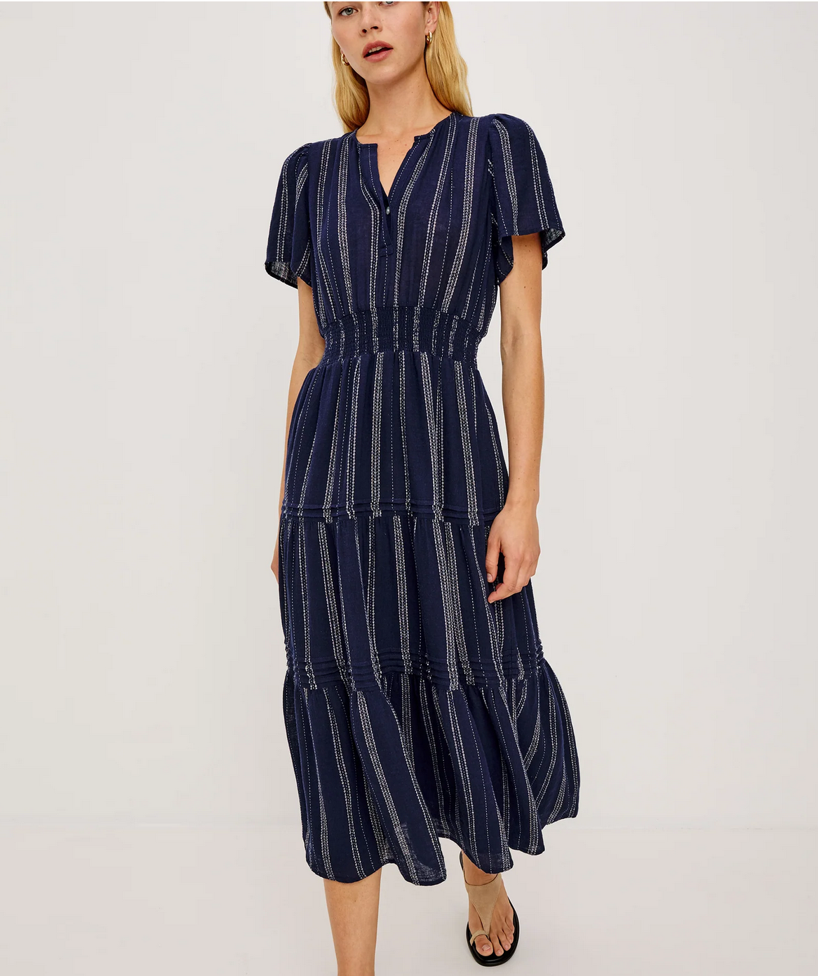 A woman wears the Rails Cannes Dress - Blue, a navy short-sleeved linen midi dress with vertical white stripes, a smocked waist, and tiered skirt, standing against a plain background.