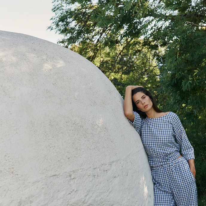 A woman wearing the Indi & Cold Multi-position Wrap Top in blue gingham leans against a large white dome outdoors, with trees behind her.