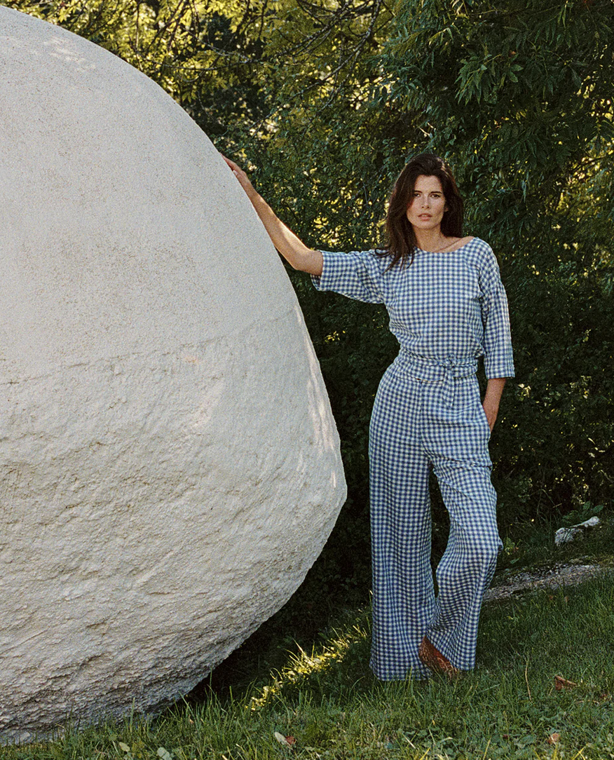 A woman wears the Indi & Cold Multi-position Wrap Top in blue gingham, standing outdoors by a large round stone structure with greenery in the background.