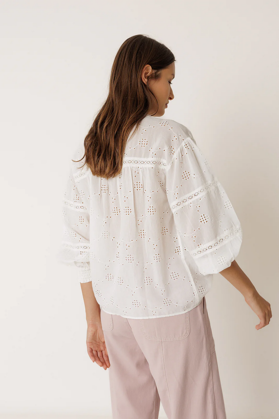 Woman wearing a white embroidered blouse and light pink pants on a plain background