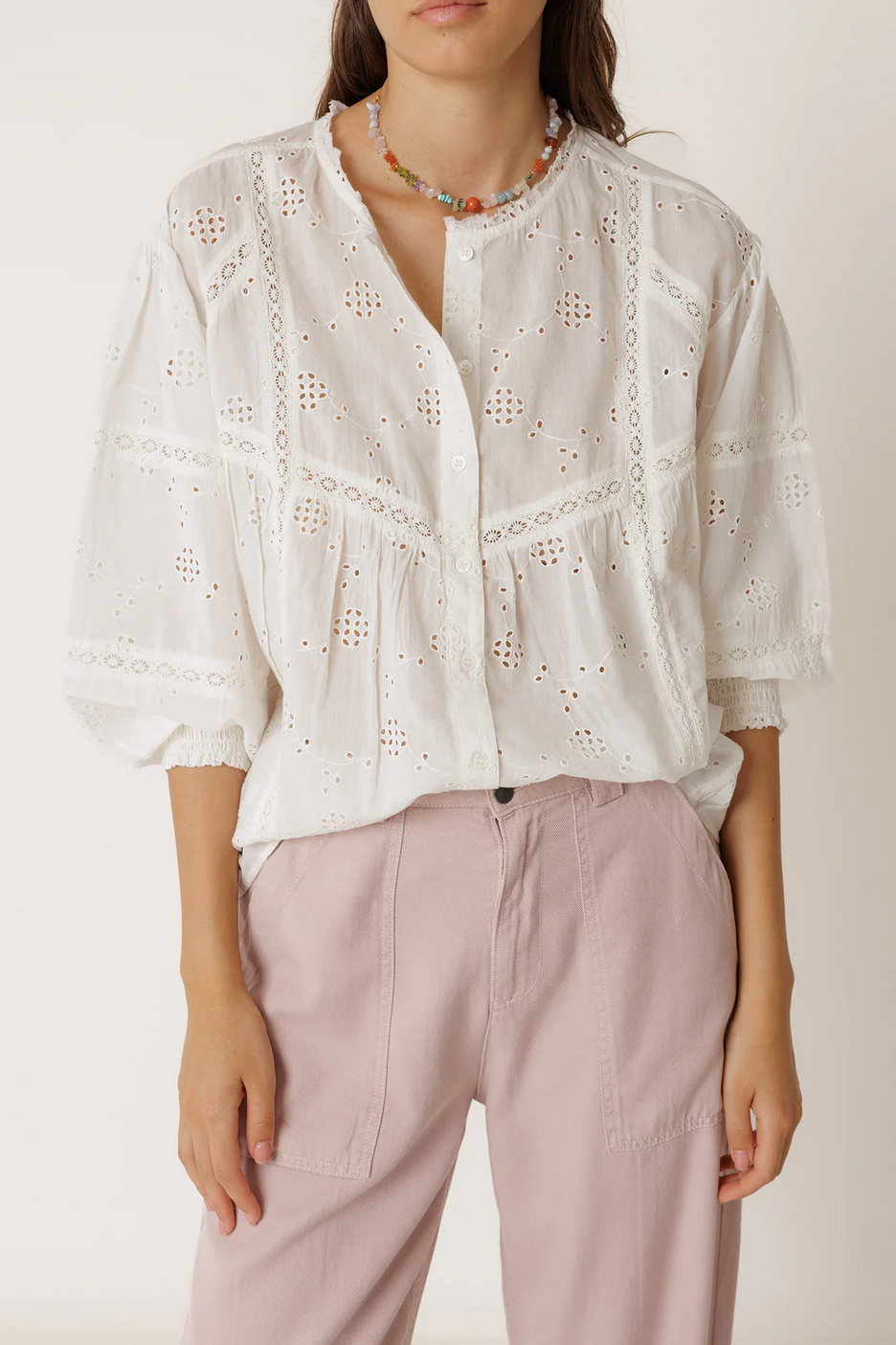 Woman wearing a white lace blouse and pink pants against a plain background