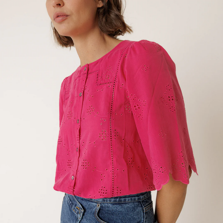 Person wearing a bright pink blouse by indi and cold with cut-out details and blue jeans on a plain background