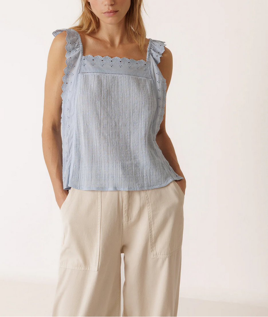 Woman wearing a light blue sleeveless top with ruffled details and beige pants, standing against a white background with 'INDI&COLD' branding.