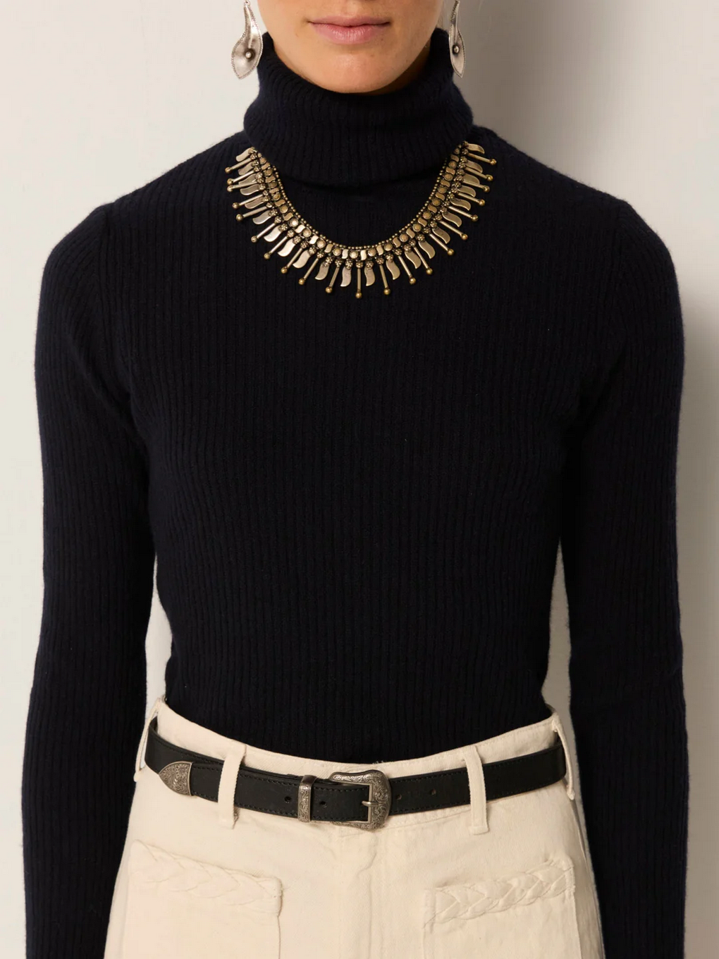 Person wearing a navy ribbed knit Maison Hotel Dolcevita Sweater, a gold statement necklace, cream high-waisted pants, and a black belt with a decorative buckle.