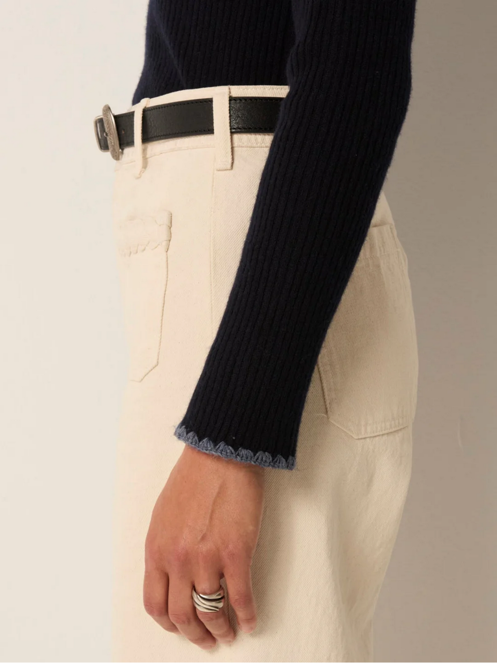 A person wearing the Maison Hotel Dolcevita Sweater in navy, paired with cream high-waisted pants, a black belt, and silver rings on their hand.