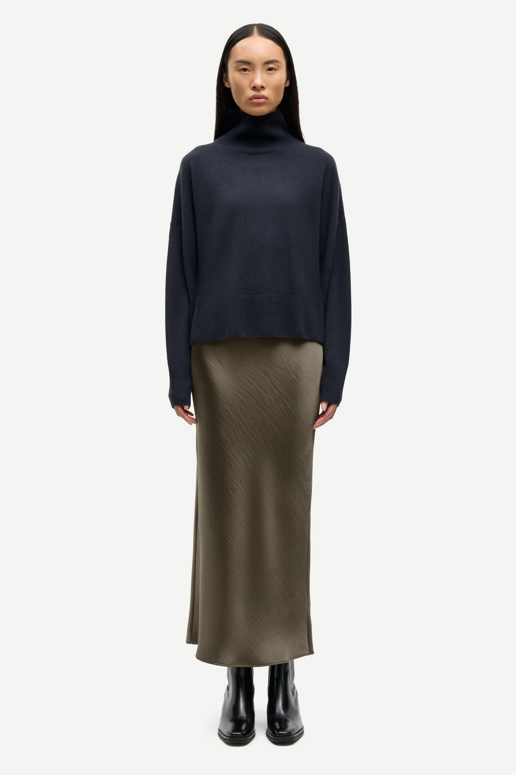 A person faces forward in a black turtleneck, the Samsøe Samsøe Saagneta Long Satin Skirt in Deep Forest, and black ankle boots, posed against a plain white background.