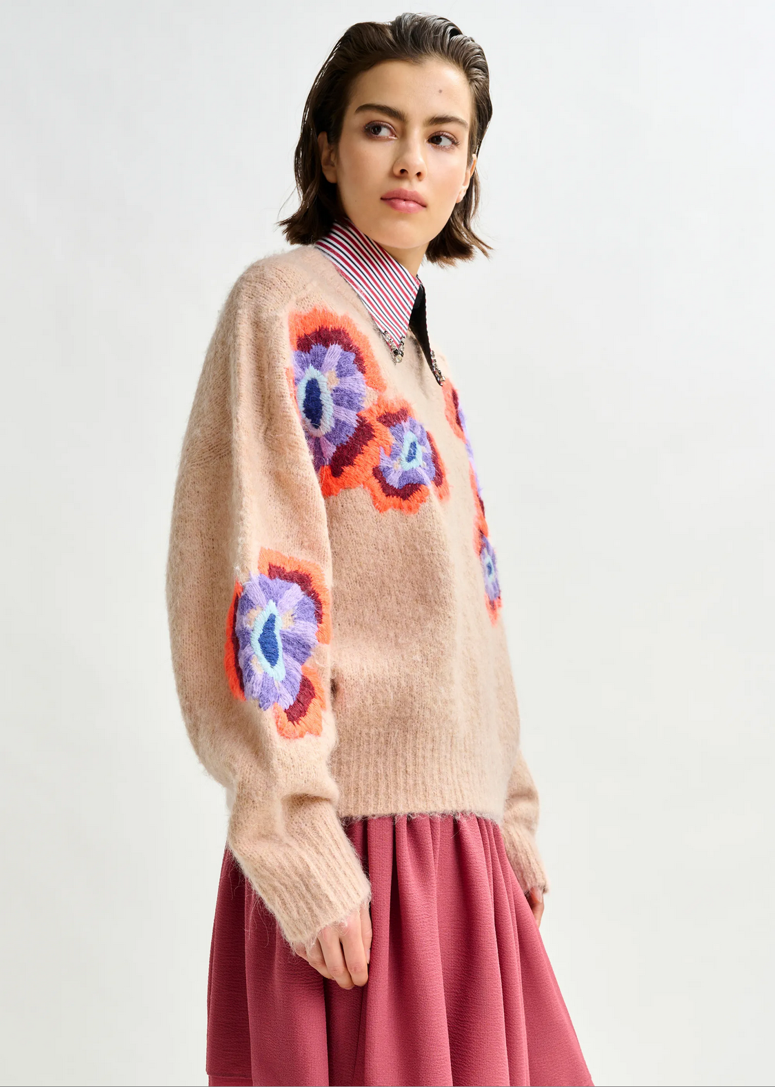 A person wears Essentiel Antwerp’s Impossible Jacquared Knit in beige—its vivid purple and orange floral design stands out atop a striped shirt and pink skirt, blending cozy flair with bold style against a plain background.