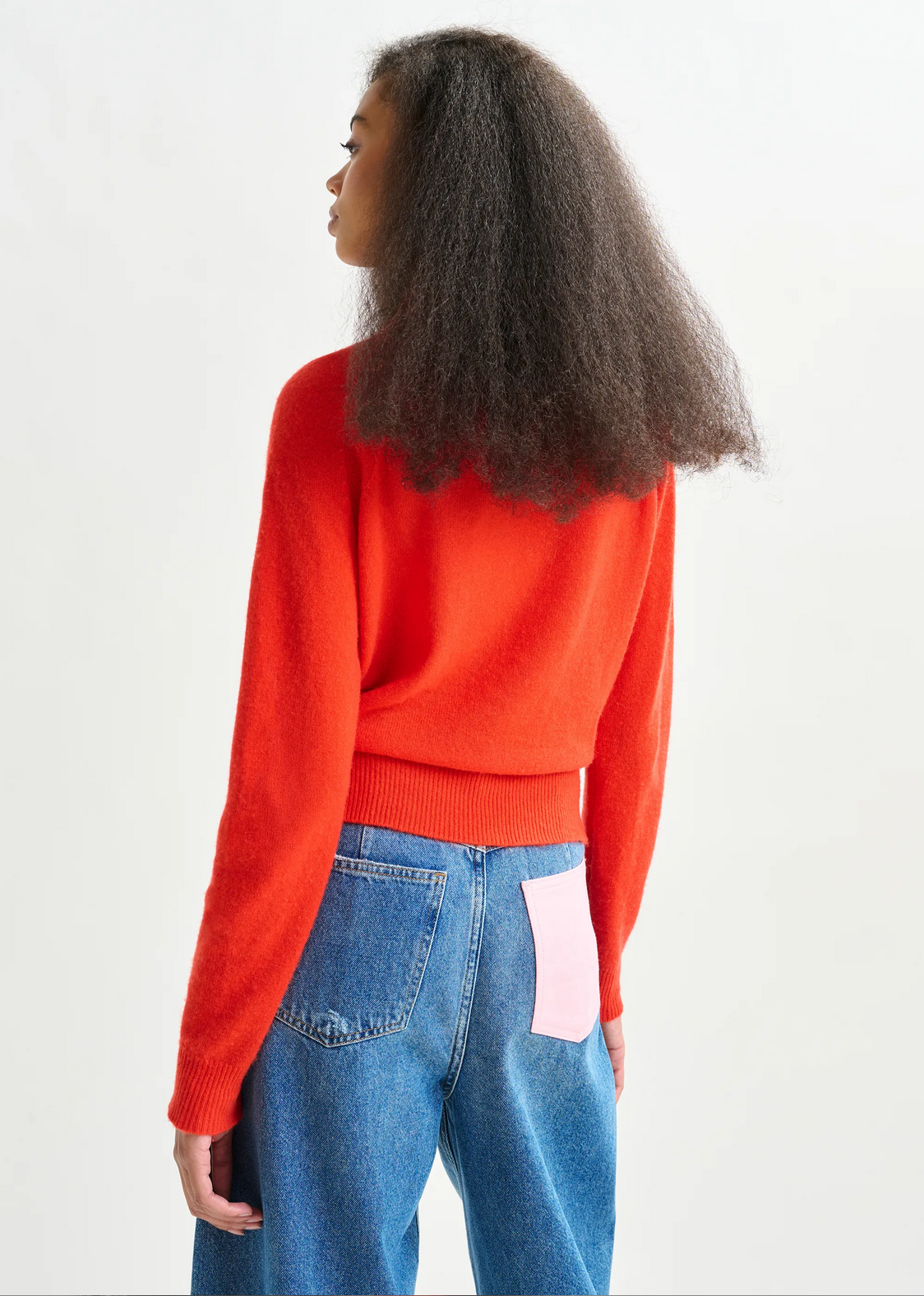 A person with long curly hair wears the Essentiel Antwerp Invited Cashmere Sweater in orange and blue jeans with a pink patch on the back pocket, standing against a plain light background.