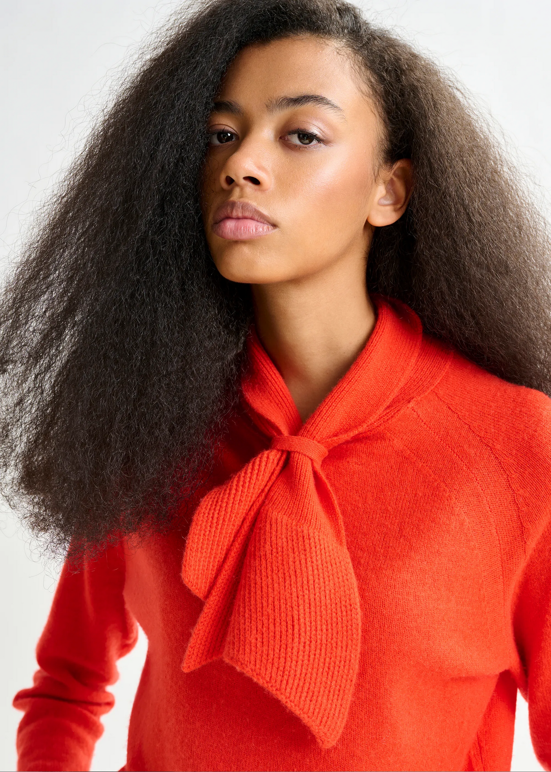 A woman with long, voluminous hair showcases the Invited Cashmere Sweater in orange by Essentiel Antwerp, featuring a statement knotted bow-tie collar detail, posed against a neutral background.