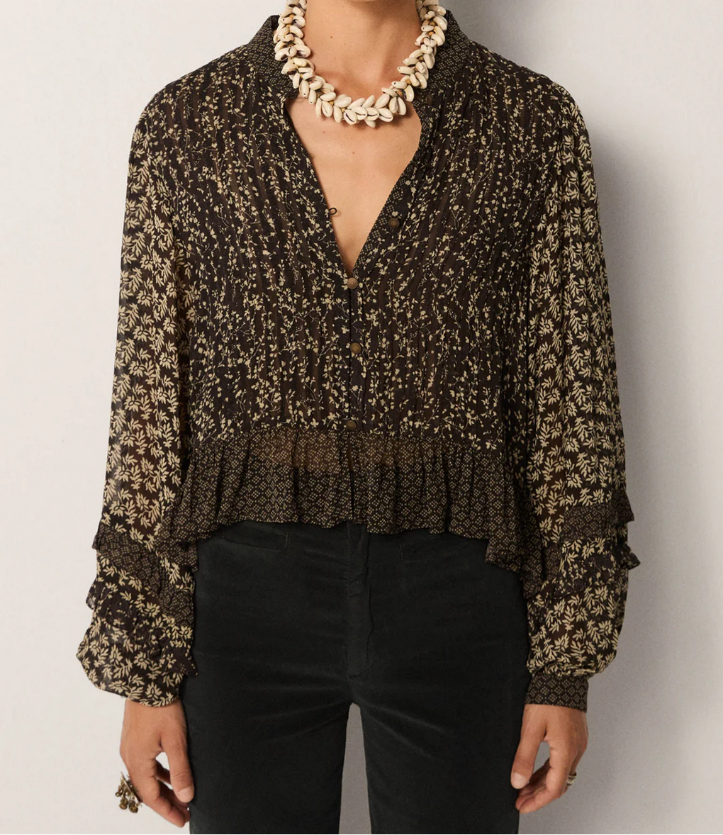 Person wearing the Maison Hotel Catalina Blouse – Black Sheep, featuring a dark pattern, billowing sleeves, and a ruffled hem, paired with black pants and a chunky shell necklace.