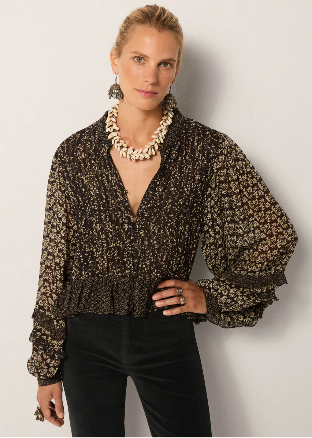 A person wears the Maison Hotel Catalina Blouse – Black Sheep with billowing sleeves, black pants, a shell necklace, large earrings, and rings, standing against a plain light backdrop.
