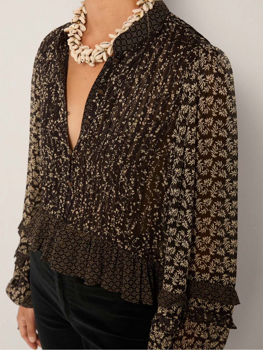 A person wears the Maison Hotel Catalina Blouse – Black Sheep, featuring dark floral patterns and billowing sleeves, paired with a chunky white seashell necklace.