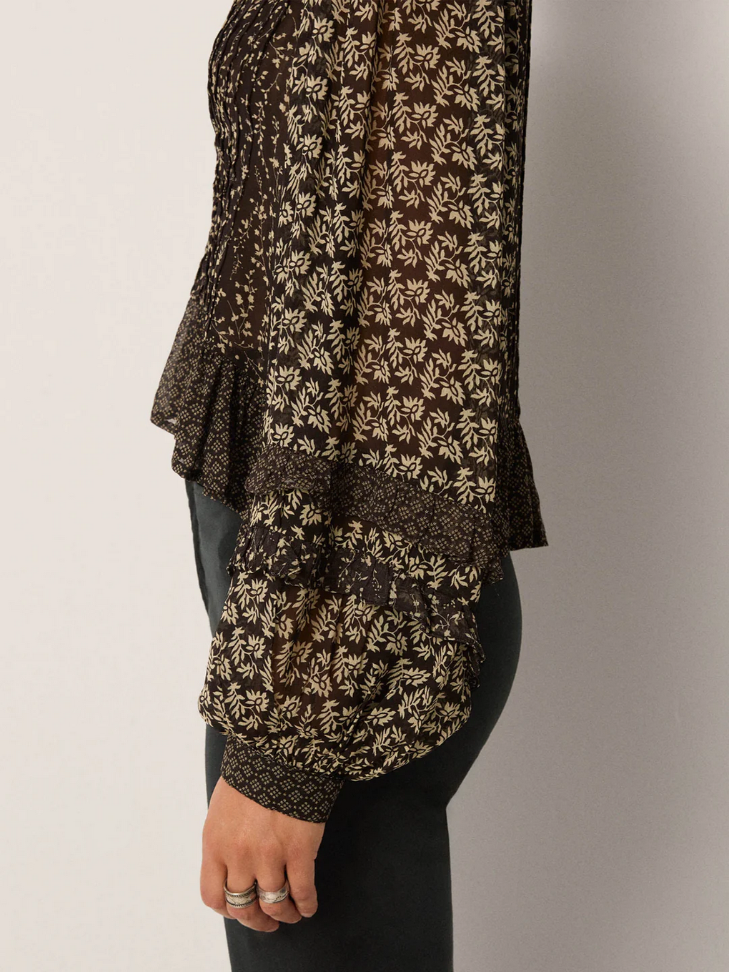 A person is shown from the side against a plain background wearing Maison Hotel’s Catalina Blouse – Black Sheep, a long-sleeved sheer top with billowing sleeves and a floral pattern, paired with dark pants.