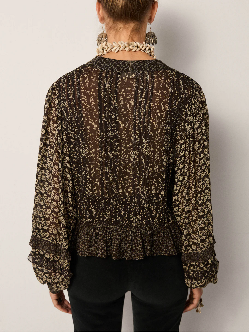A woman is shown from the back wearing a sheer, patterned Maison Hotel Catalina Blouse – Black Sheep with billowing sleeves, paired with black pants and a chunky shell necklace.